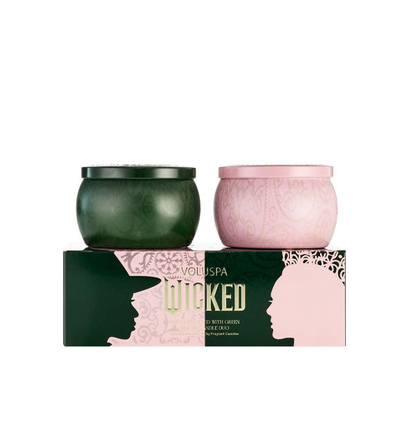 Two patterned candle tins with lids, one green and one pink, atop a Wicked-themed gift box by Voluspa printed with silhouetted profiles of both Elphaba and Glinda