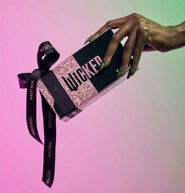 Model's hand with long, shimmery, green fingernails holds a Wicked-themed Voluspa candle set gift box wrapped with a black branded bow against a pink and green backdrop