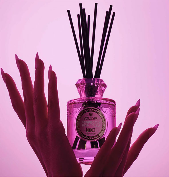 Silhouetted hands with long fingernails hold an embossed pink glass Wicked Perfectly Pink Yuzu Voluspa reed diffuser