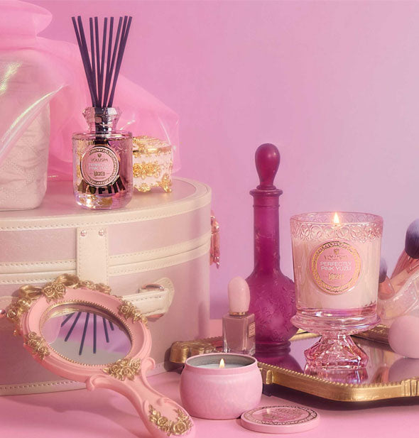 Wicked-themed Perfectly Pink Yuzu Voluspa reed diffuser, mini tin candle, and embossed glass jar candle staged with ornate hand mirror, pink decanter, and other items on a pink backdrop