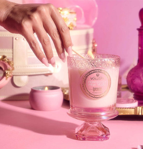 Model's hand lights a Wicked-edition Voluspa Perfectly Pink Yuzu candle in an embossed pink glass jar resting on its removed pedestal lid against a backdrop with other pink items