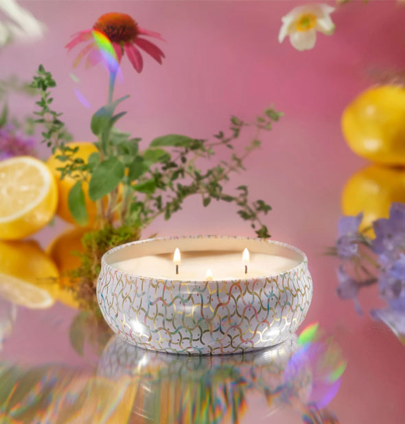 Lit 3-wick candle in decorative tin on a reflective surface with wildflowers and lemons behind it against a pink backdrop