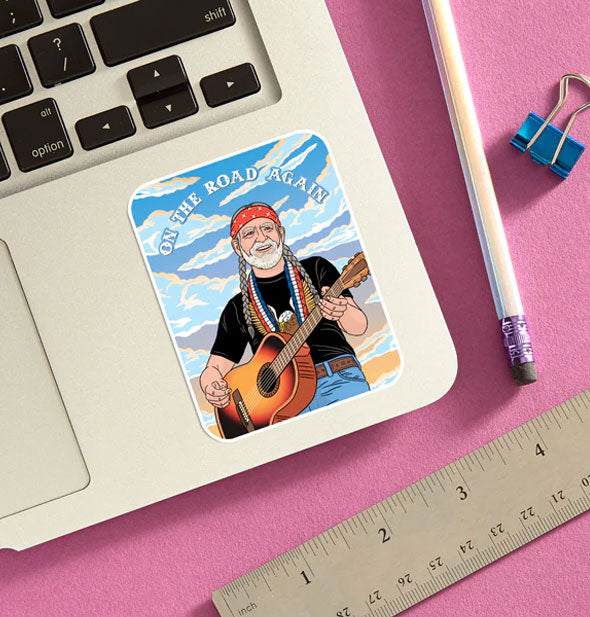 Willie Nelson "On the Road Again" sticker is applied to a laptop base panel and staged with pencil, ruler, and blue binder clip on a pink surface