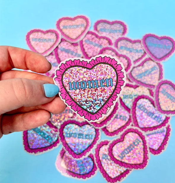 Model's hand holds a heart-shaped sticker with glittery center that says, "Women" in blue Gothic style lettering and features an outer ruffly pink border in front of a pile of others like it in the background