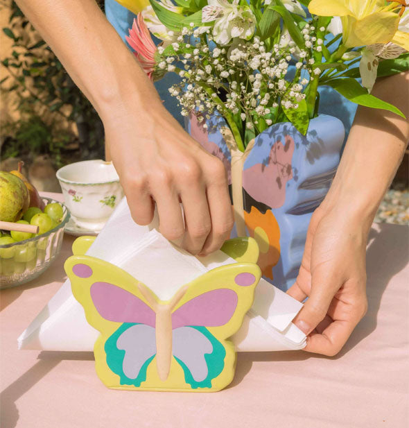 Model adjusts the napkins in a Woodland Butterfly Napkin Holder on a pink cloth-covered table set with other accoutrements
