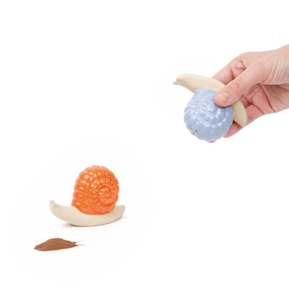 Model holds a blue-shelled ceramic snail shaker upside down near an orange-shelled ceramic snail shaker with a brown spice pile in front of it