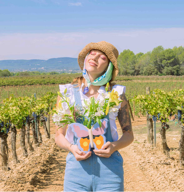 Model standing in a vineyard basking in sunlight holds a butterfly vase in both hands that has been filled with lush florals