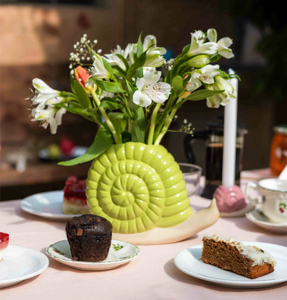 Snail vase on a tabletop with plated cake slices holds a bouquet of fresh flowers