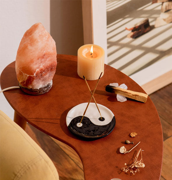 Black and white yin yang incense dish on a curved wooden tabletop staged with salt crystal lamp, burning pillar candle, palo santo stick, and dried flowers holds two burning incense sticks