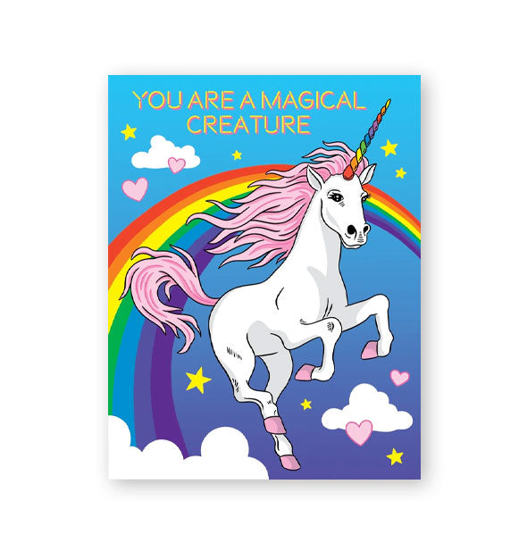 Greeting card featuring full-color illustration of a white unicorn with pink mane, tail, and hooves flying in the sky among white clouds, a rainbow, yellow stars, and pink hearts says, "You are a magical creature" at the top in yellow lettering