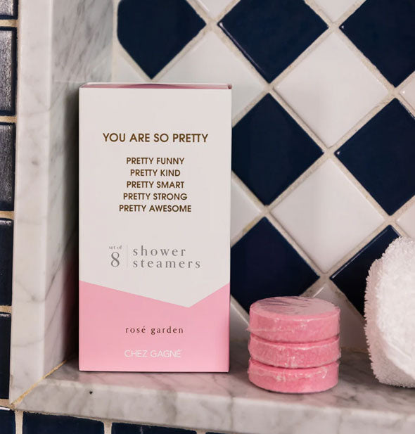 You Are So Pretty shower steamers box and three pink steamers rest on a marble bath ledge next to a rolled-up white washcloth and in front of black and white tile
