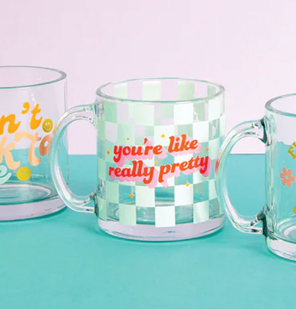 You're Like Really Pretty glass mug staged with two other designs on a teal surface against a light pink backdrop