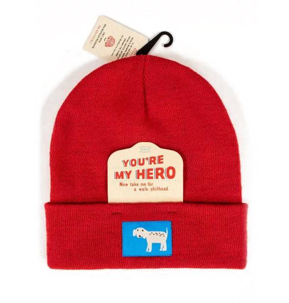 Red knit beanie with blue tag that features a white dog with black spots has a tag slipped under the brim that says, "You're my hero, now take e for a walk shithead"
