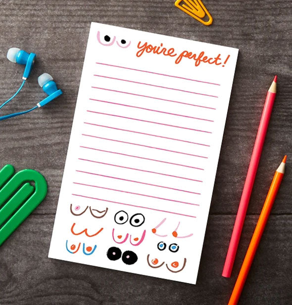 You're Perfect! boobies notepad rests on a wooden surface with green and yellow paper clips, blue earbuds, and red and orange colored pencils