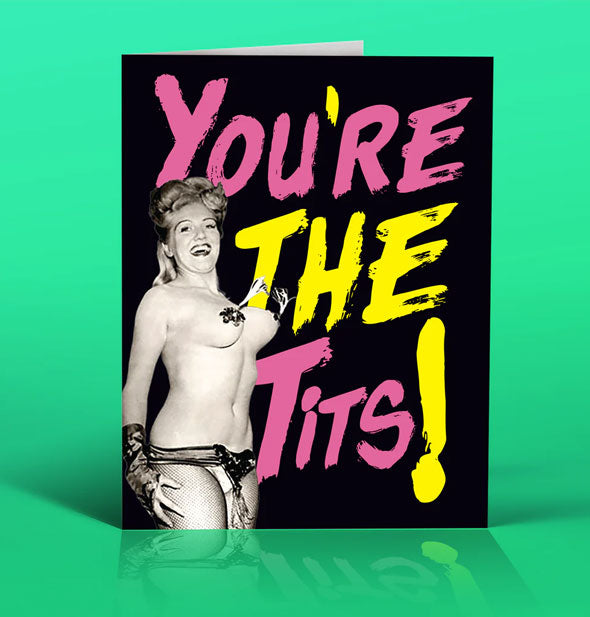 Black greeting card on green backdrop features black and white photograph of a smiling woman wearing pasties with tassels beside the words, "You're the tits!" in large pink and yellow lettering