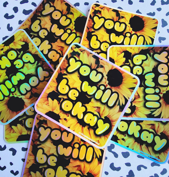 Pile of square stickers on a black and white speckled surface feature a sunflower design with the words, "You will be okay" overtop in yellow bubble lettering