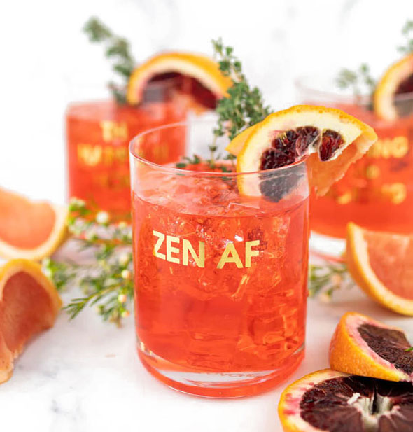 Zen AF rocks glass holds a reddish-orange iced beverage garnished with citrus slices and fresh herb sprigs