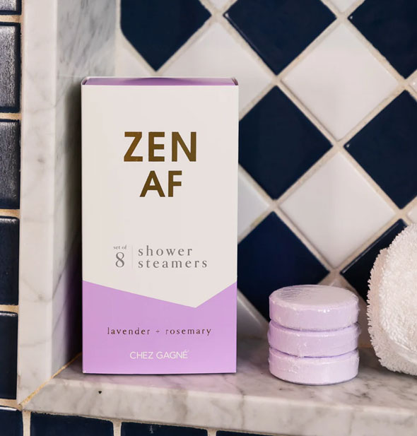 Zen AF shower steamers box and three light purple steamer discs rest on a marble bath ledge next to a rolled-up white washcloth in front of black and white tile