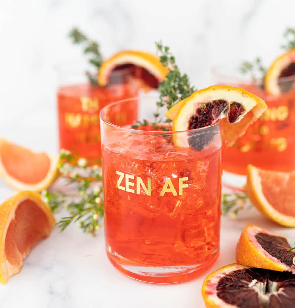 Zen AF rocks glass holds a reddish-orange iced beverage garnished with citrus slices and fresh herb sprigs