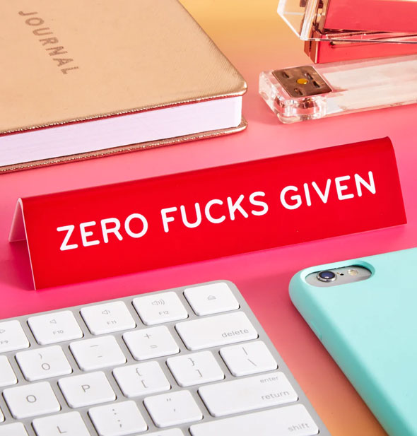 Red "Zero Fucks Given" desk sign is staged with a computer keyboard, smartphone, gold journal, and stapler on a pink surface