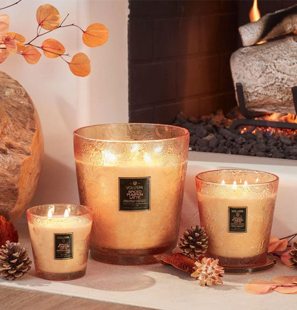 Three orange embossed glass jar candles with pine cones on a lit stone hearth