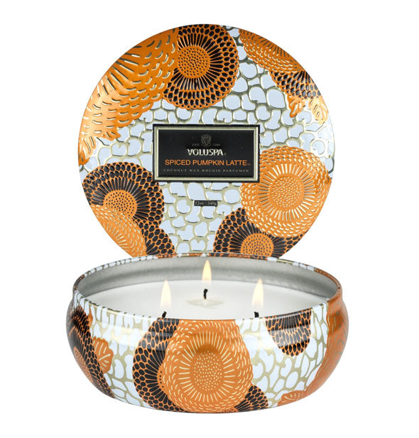3-wick candle in a flat, rounded tin with black, orange, white, and metallic design accents and a matching lid standing up in the background.