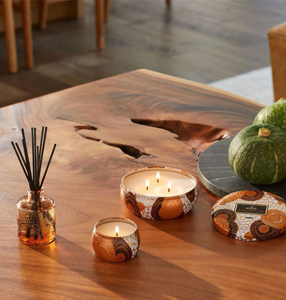 Decorative brown, white, and orange candle tins with orange embossed glass reed diffuser on a wooden tabletop
