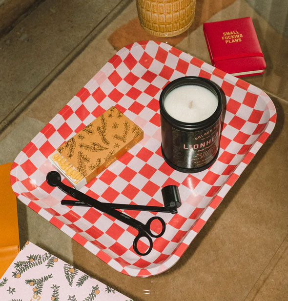 Red and pink checkered tray rests on an upholstered surface and is staged with candle, matches, and other accoutrements