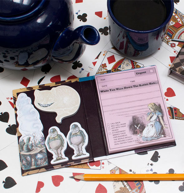 Open Wonderland Sticky Notes book shows pad and sample notes inside including the Cheshire Cat and Tweedledee and Tweedledum and is staged with playing cards and a dark blue tea set