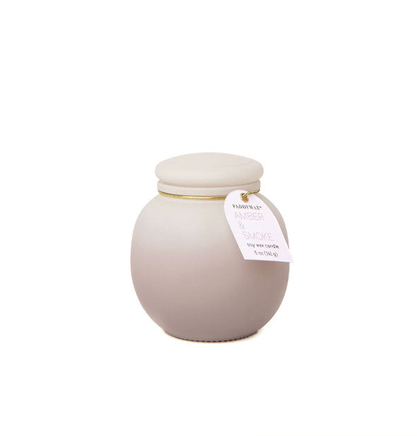 Rounded candle jar with frosted matte finish and beige ombre coloration is topped with a lid and tag on a gold band