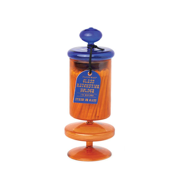 Amber glass bottle filled with matchsticks and topped with a knobbed blue lid features a blue hang tag that reads, "Glass matchstick holder, 140 matches, strike on glass"