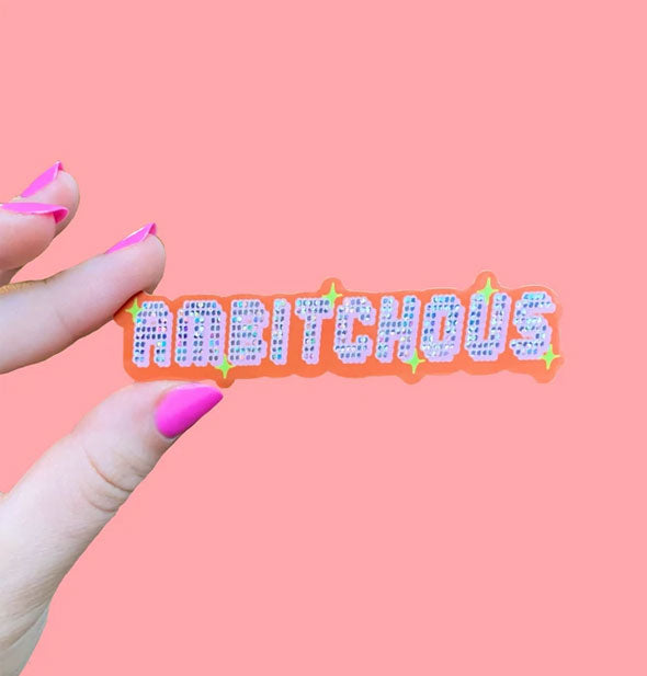Model's hand holds a sticker that says, "Ambitchous" in digital-style lettering with glitter and star accents