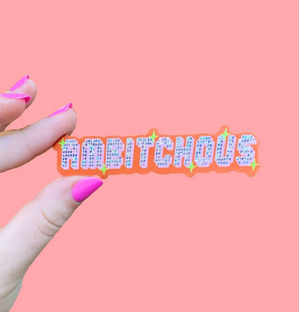 Model's hand holds a sticker that says, "Ambitchous" in digital-style lettering with glitter and star accents