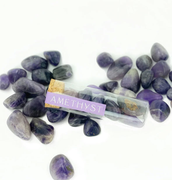 Clear vial of polished purple amethyst stones with cork cap and printed purple label rests on top of other loose amethyst stones