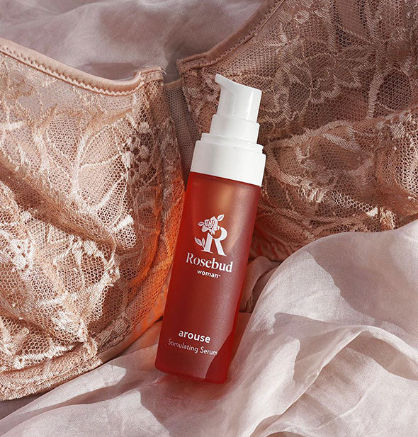 Coral and white bottle of Rosebud Woman Arouse Stimulating Serum rests on a taupe lace brassiere and white chiffon