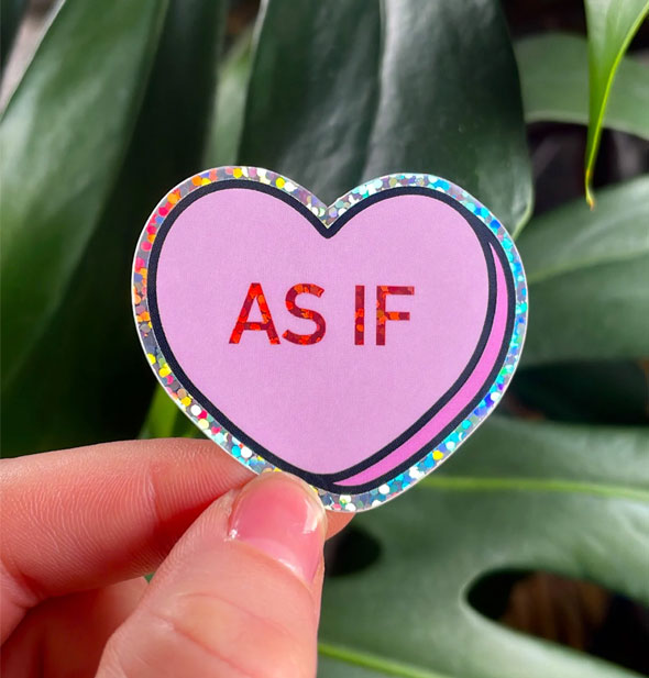 Model holds a heart-shaped sticker in hand with a rainbow holographic border and "As If" printed in the center in holographic red lettering; green monstera leaves are in the background