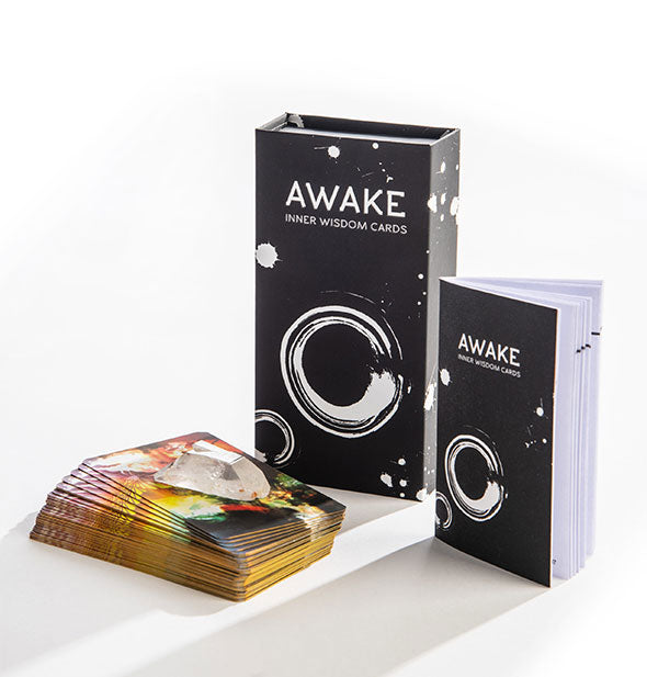 A rectangular black box with white graphics and the words, "Awake Inner Wisdom Cards" sits alongside a booklet of the same design and a colorful card deck with clear crystal on top.