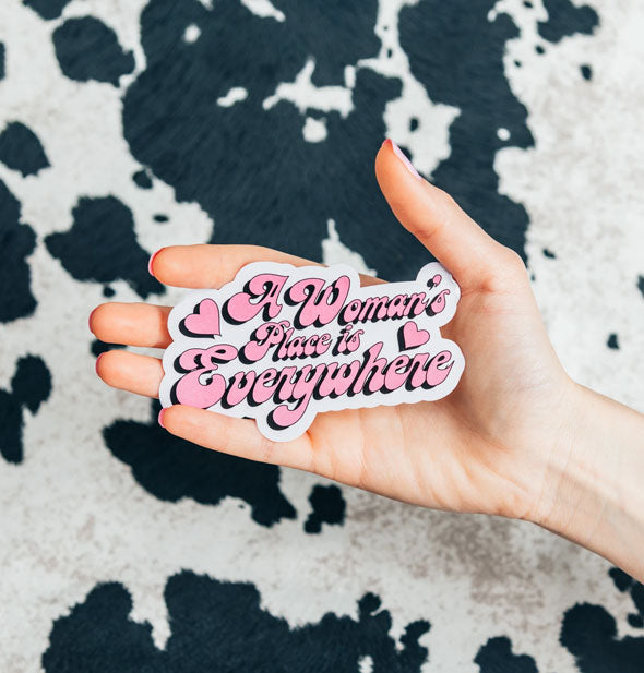 Model's hand holds A Woman's Place is Everywhere sticker against a cowhide print backdrop