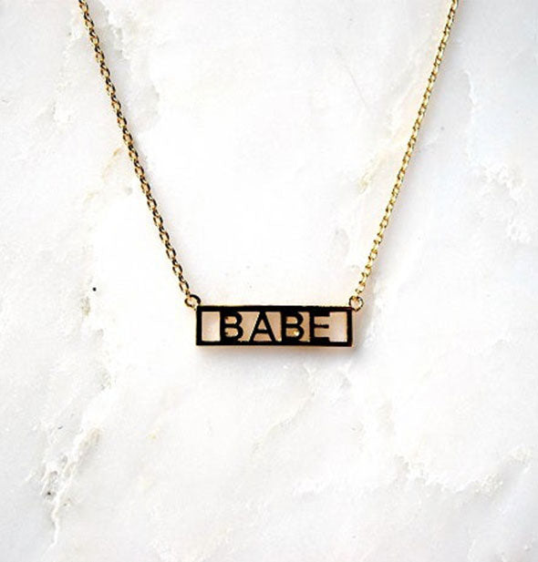 Gold bar necklace on white marble surface says, "Babe"