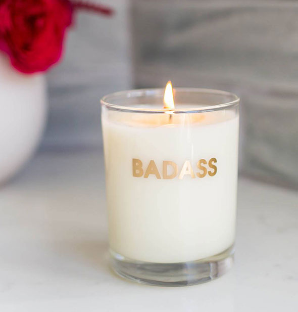 Lit candle in glass vessel printed with "Badass" in metallic gold lettering sits on a marble backdrop with vase of red roses in the backround