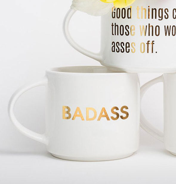 White mug with gold foil "Badass" imprint is displayed with other mugs.