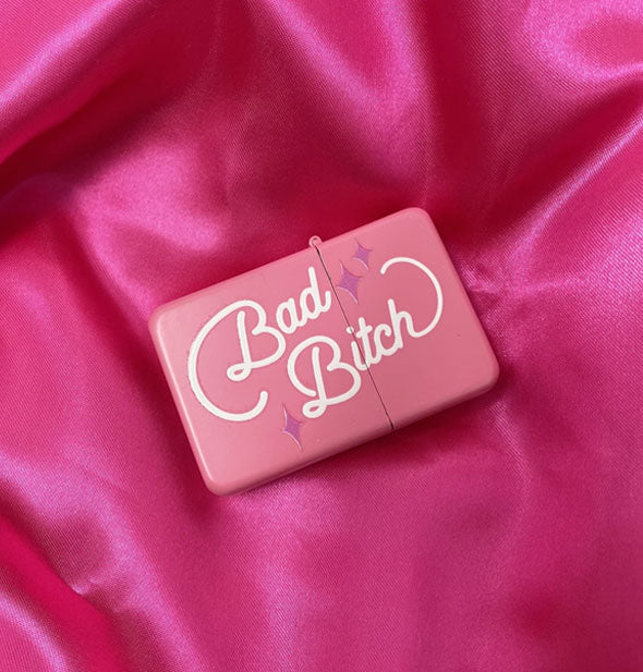Pink lighter on pink satin says, "Bad Bitch" in white script with purple star accents