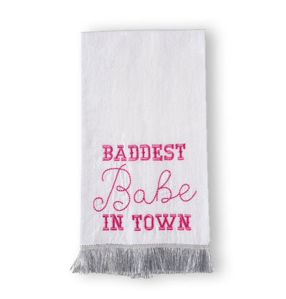 Folded white tea towel with silver fringe at the bottom says, "Baddest Babe in Town" in hot pink embroidery