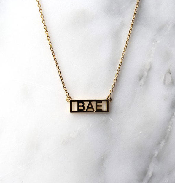 Gold bar necklace on white marble surface says, "Bae"