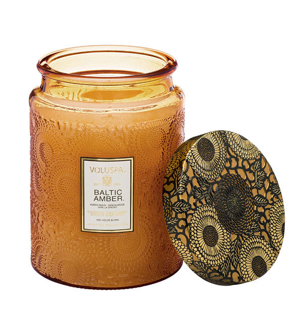 Golden embossed glass Baltic Amber Voluspa candle jar with black and gold metallic floral lid set to the side