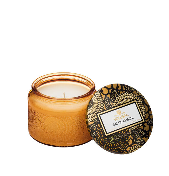 Golden embossed glass Baltic Amber Voluspa candle jar with black and gold metallic floral lid set to the side
