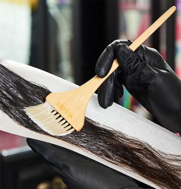 A stylist applies hair color to a section of a client's hair with a bamboo ColorTrak brush