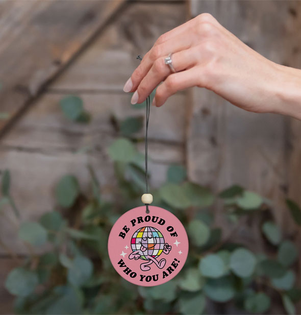 Model's hand holds a round pink car air freshener featuring a colorful disco ball graphic and the words, "Be proud of who you are!" from its string against a botanical and wood backdrop