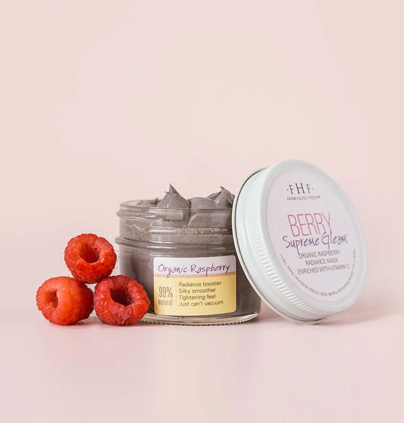Opened pot of Berry Supreme Gleam Organic Raspberry Radiance Mask with lid propped up and to the side is staged with three red raspberries at left