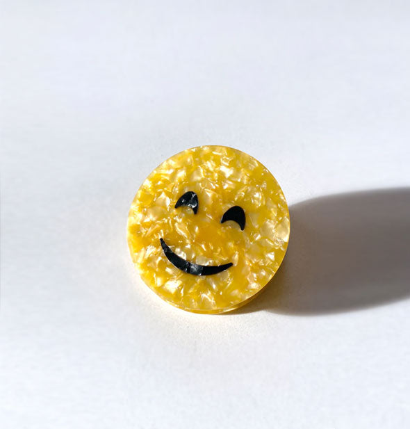 Round yellow quartz-effect hair clip with black smiley face design casts a harsh shadow on a light-colored surface